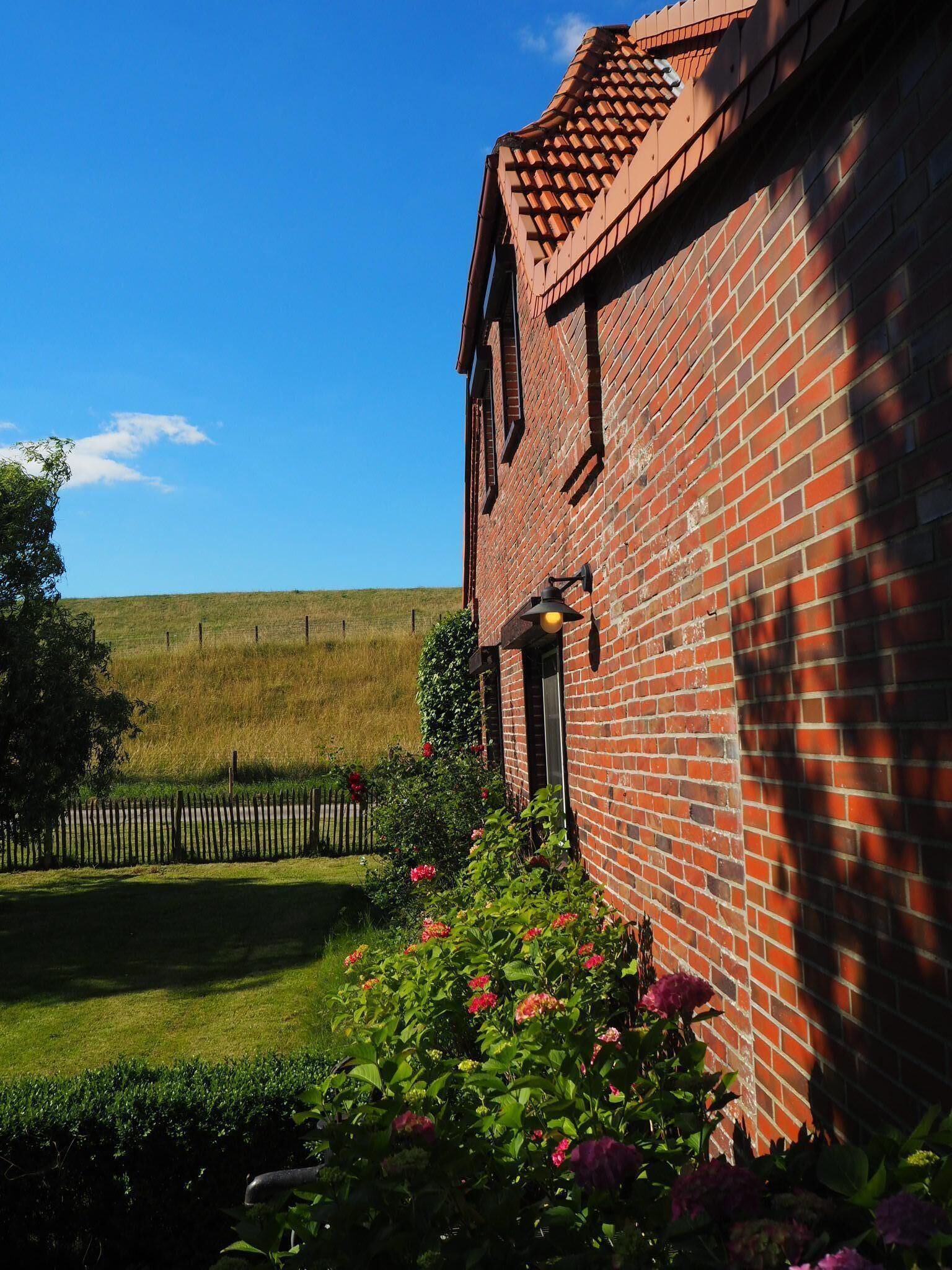 A brick building with a red-tiled roof, a small porch with a lamp, and a well-maintained garden with flowers and a fence.
