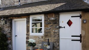 A stone cottage with a white door, a window with a red curtain, and a table with a bottle and glasses.