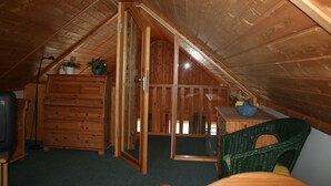 A cozy attic bedroom with wooden walls and a sloped ceiling. It features a wooden dresser, a green wicker chair, and a small table with a vase. - The house is located right on the edge of the Müritz National Park (Userin)