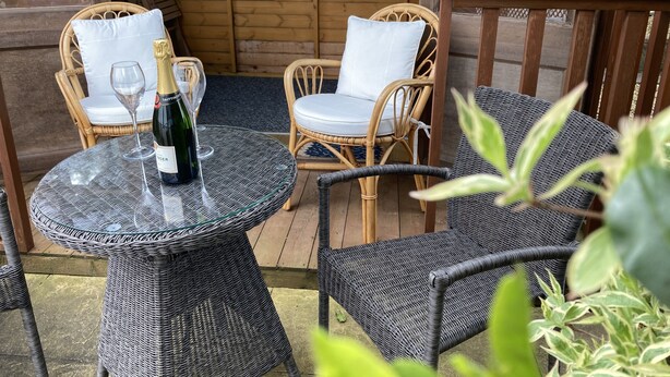 Ein Korbtisch mit einer Champagnerflasche und einem Glas, zwei Korbstühle mit weißen Kissen sowie eine Holzwand im Hintergrund.