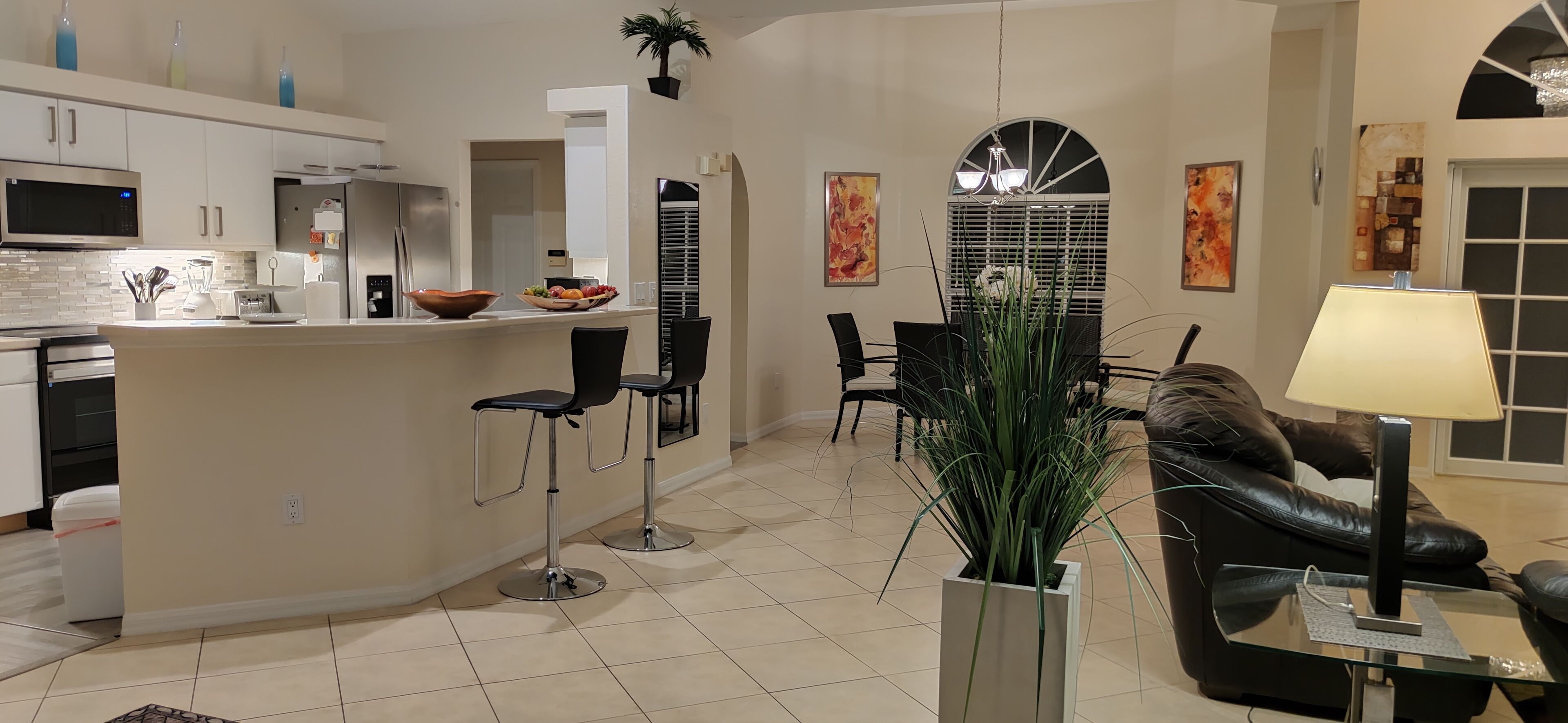 A modern kitchen with a microwave, refrigerator, and a fruit bowl on the counter. There are two black chairs and a dining table with a plant in the center. A black leather sofa and a glass coffee table are visible in the living area.