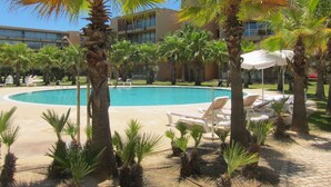 A pool area with lounge chairs and umbrellas, surrounded by palm trees and a building in the background.