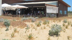 A modern building with a covered outdoor seating area and a sandy foreground with sparse vegetation.