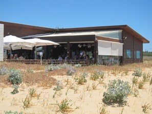 Un edificio moderno con un área de estar al aire libre cubierta y un primer plano arenoso con vegetación escasa.