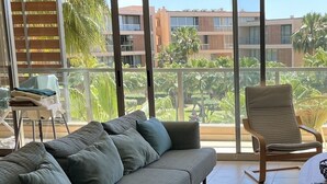 A modern living room with a grey sectional sofa, a round coffee table, and a lounge chair. The room has large windows offering a view of a residential area with palm trees and buildings.
