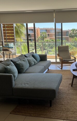 Una sala de estar moderna con un sofá seccional gris, una mesa redonda y un sillón. La habitación tiene ventanas grandes que ofrecen vista a una zona residencial con palmeras y edificios.