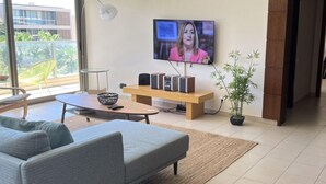 A modern living room with a grey sofa, a coffee table, and a TV mounted on the wall. There is a wooden bench and a potted plant in the corner.
