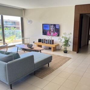 Una sala moderna con un sofá gris, una mesita de centro y un televisor montado en la pared. Hay un banco de madera y una planta en maceta en la esquina.