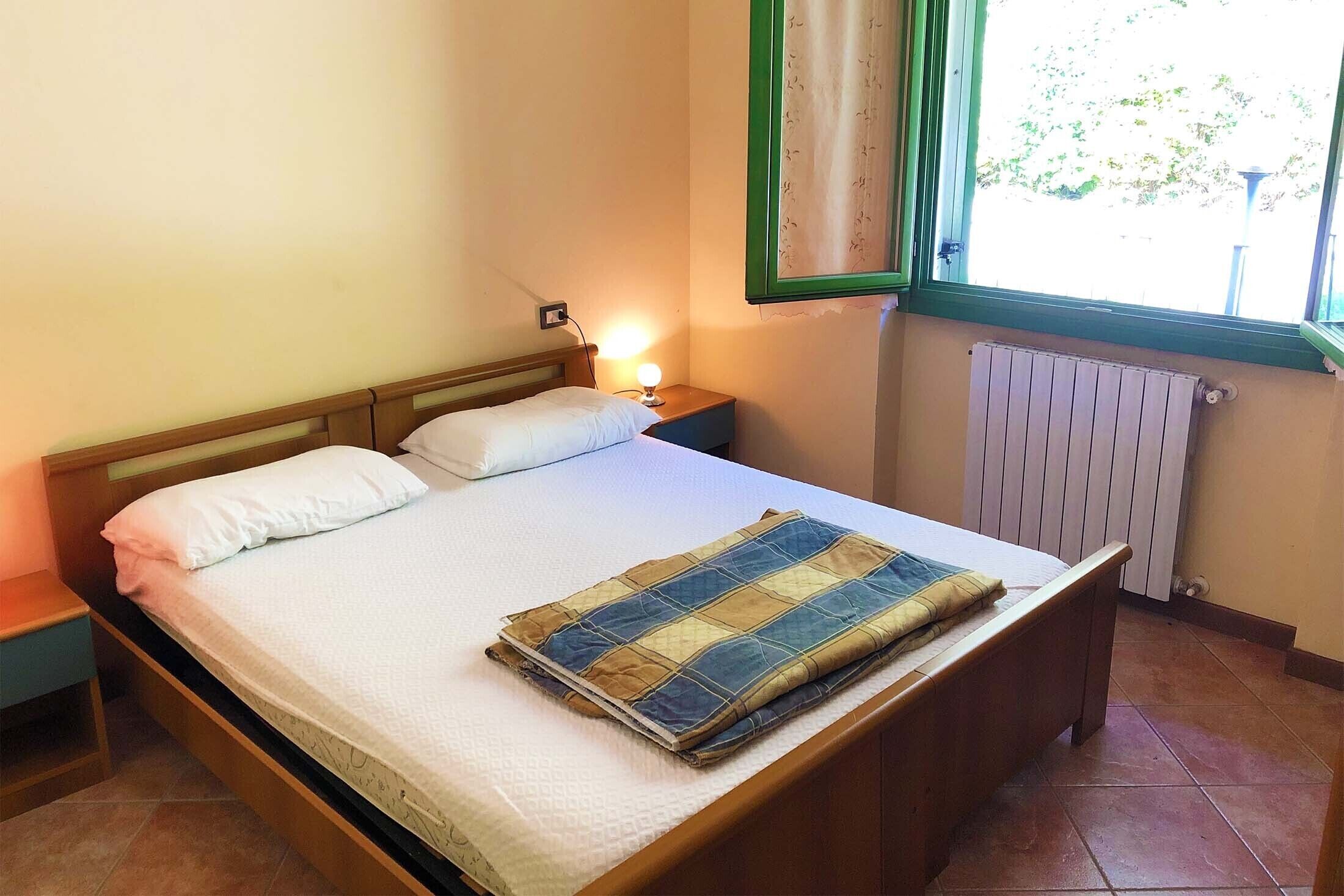 Un letto singolo con coprialetto, un comodino con una lampada, una finestra vista su del verde e un termosifone.
