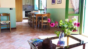 Una zona de menjador amb taula i cadires de fusta, un ram de flors i vista a la cuina a través d'una finestra.