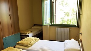 Una habitació amb dos llits, un armari de fusta, una finestra amb vista a vegetació i un radiador.