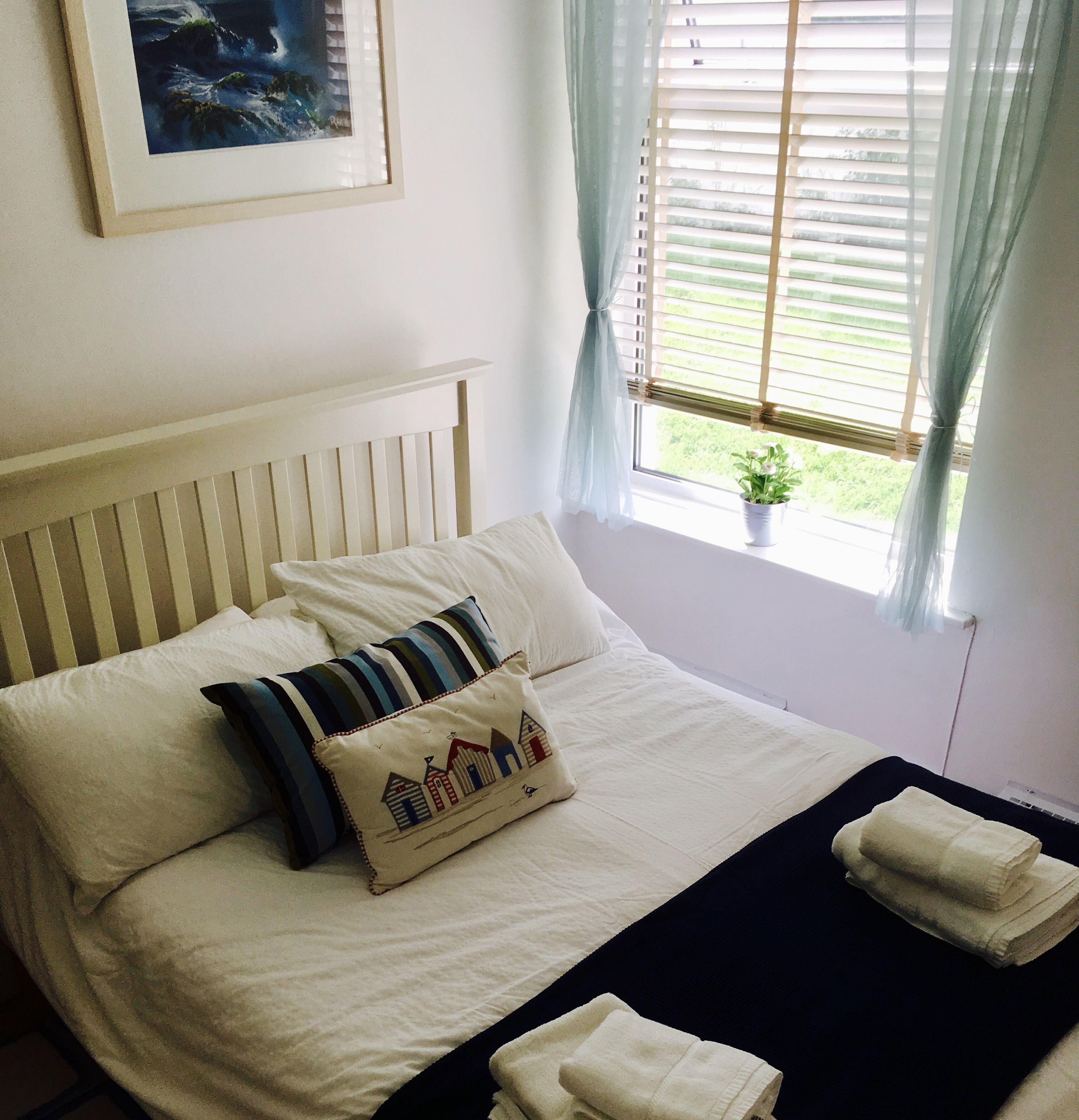 A neatly made bed with white bedding, a pillow with a house design, and a window with blinds and curtains.