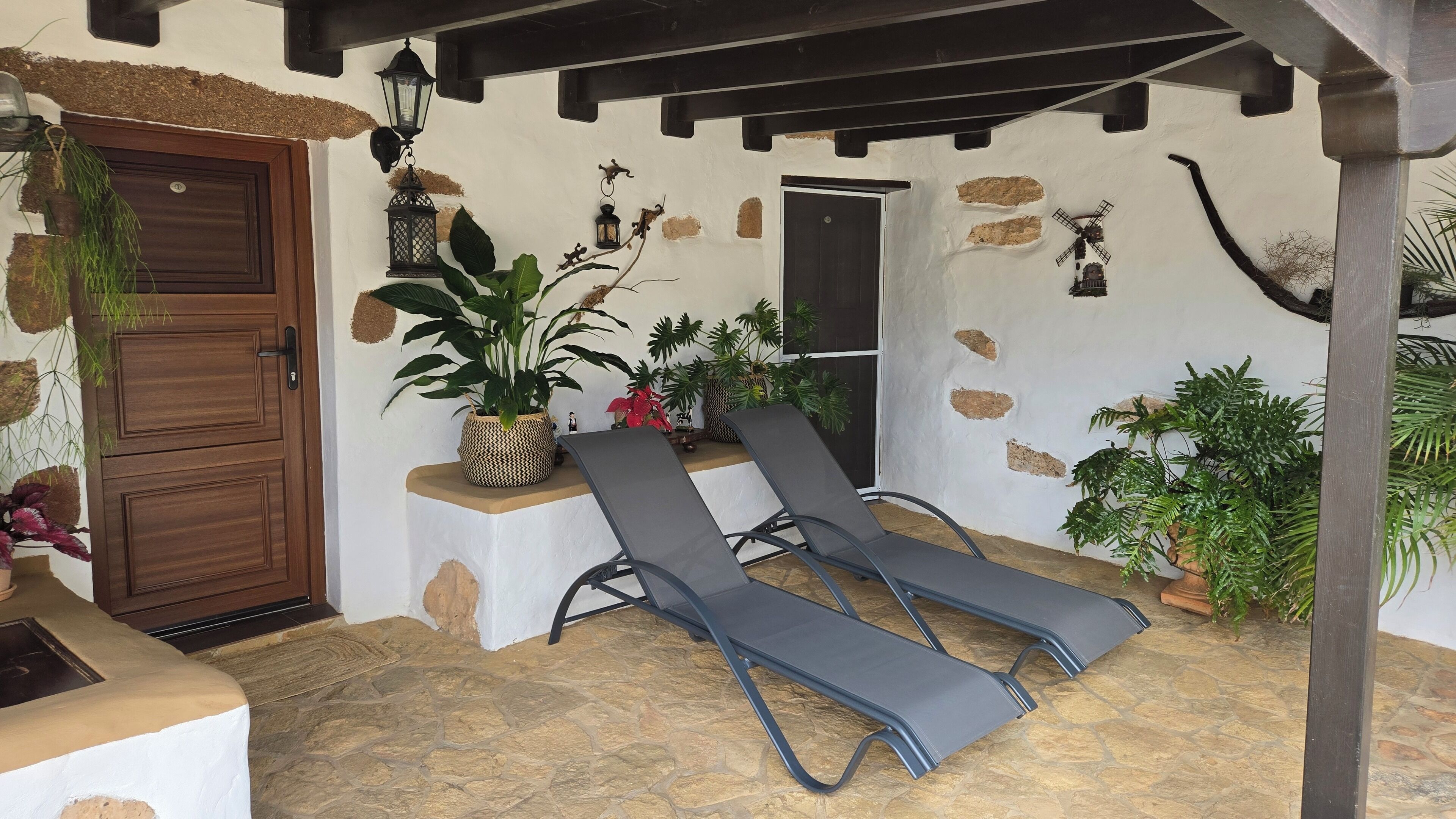 A covered outdoor seating area with two lounge chairs, a potted plant, and a door.