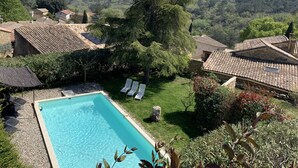 A swimming pool surrounded by a garden with lounge chairs, a stone building with a tiled roof, and a view of a forested hillside. - Character house, Mas du 17è,quiet,private pool 9x4,5m,air-conditioned,near Uzes (Le Pin)