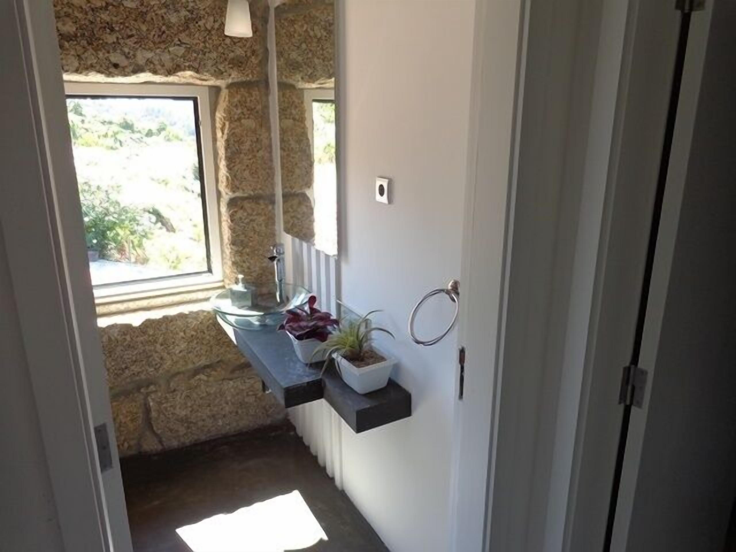 Un couloir étroit avec un mur en pierre, une fenêtre donnant sur la verdure, un lavabo en verre agrémenté d’une plante et une porte avec un porte-serviette.