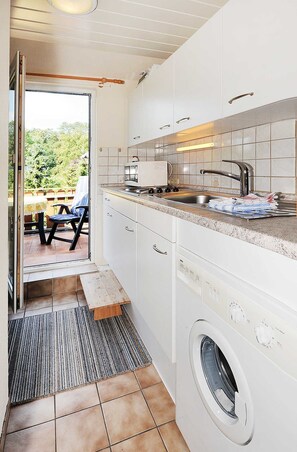 A compact kitchen with a built-in washing machine, a dining area with a table and chairs, and a view of greenery outside. - Apartment - Ferienhaus Zur Schmiede (Dunum)