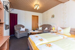 Ett hotellrum med en säng, två fåtöljer, en byrå i trä, en röd gardin, ett trätak och en vägglampa.