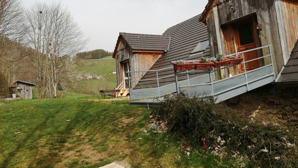 Ein rustikales Holzhaus mit geneigtem Dach, einer kleinen Veranda mit Metallgeländer und einem Gartenbereich mit Topfpflanzen.