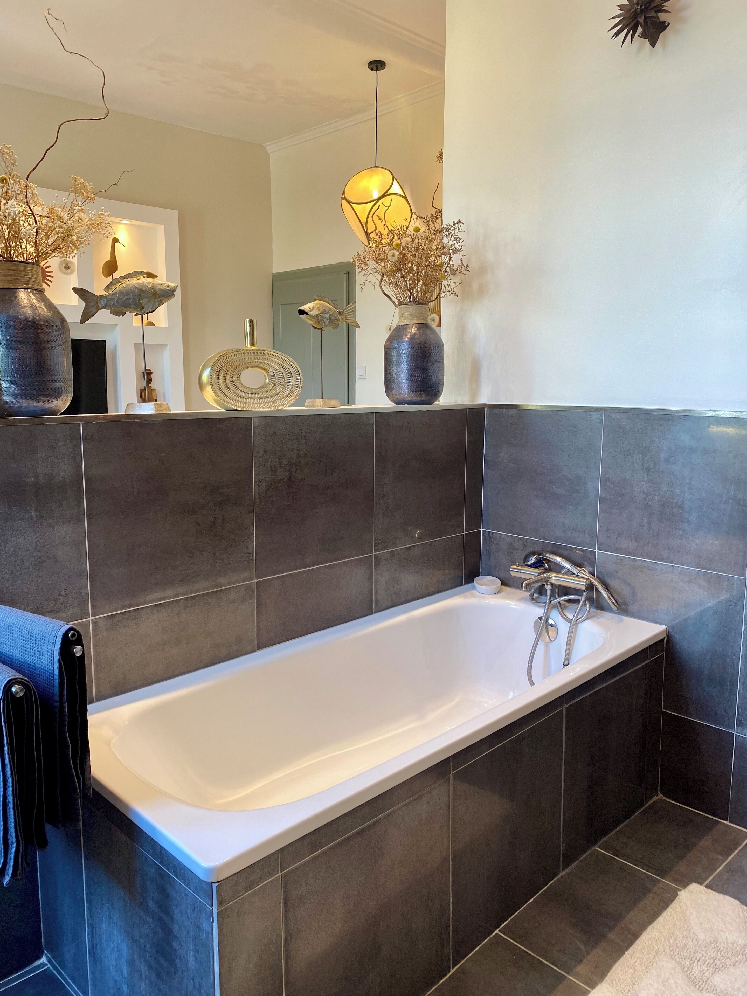 Une salle de bain moderne avec une grande baignoire, des murs recouverts de carreaux foncés et des éléments décoratifs, notamment des sculptures de poissons et des vases.