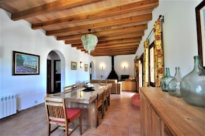 Zona pranzo con soffitto in legno, un lampadario e un lungo tavolo in legno con sedie. Sono presenti luci a parete e un camino sullo sfondo.
