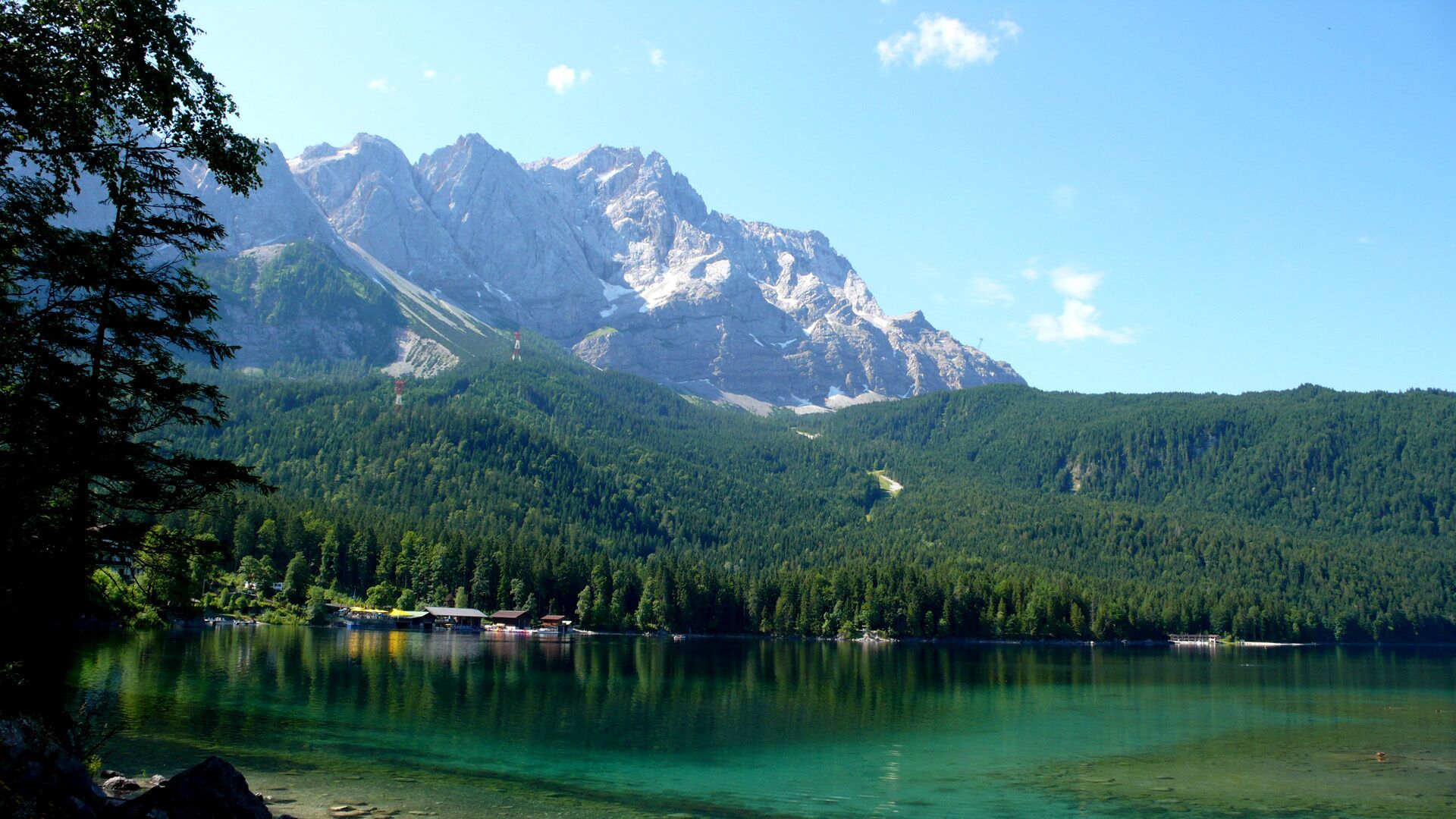 A lake with clear water surrounded by forested mountains and a few buildings on the shore.