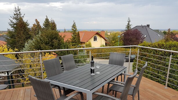 Varanda com mesa e cadeiras, rodeada por grade de metal, com vista para área residencial com casas e árvores.