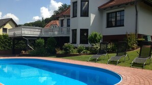 Uma piscina com borda de tijolos, cercada por um gramado bem cuidado e um edifício branco com varanda.