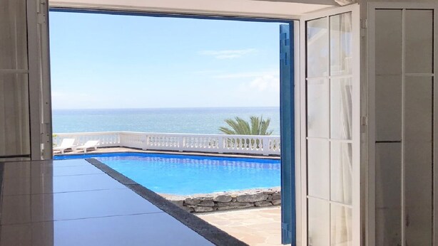 Une porte ouverte donnant sur une piscine, avec vue sur l’océan, et une clôture blanche.