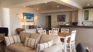 Un salon avec un canapé beige, des chaises blanches, une table à manger en bois et, au fond, un coin cuisine.