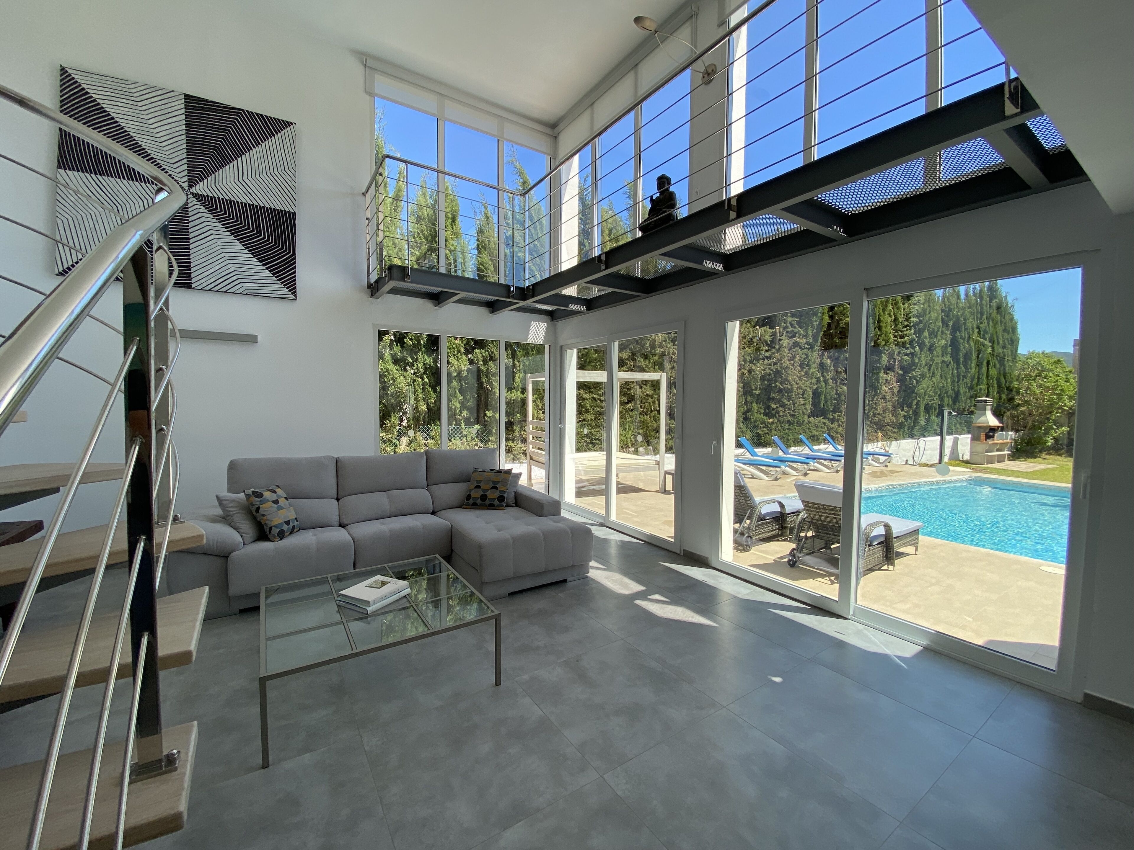 Un vaste espace de vie agrémenté d’un escalier moderne, d’un canapé d’angle gris, d’une table basse en verre et de grandes fenêtres offrant une vue sur une piscine et un espace salon extérieur.