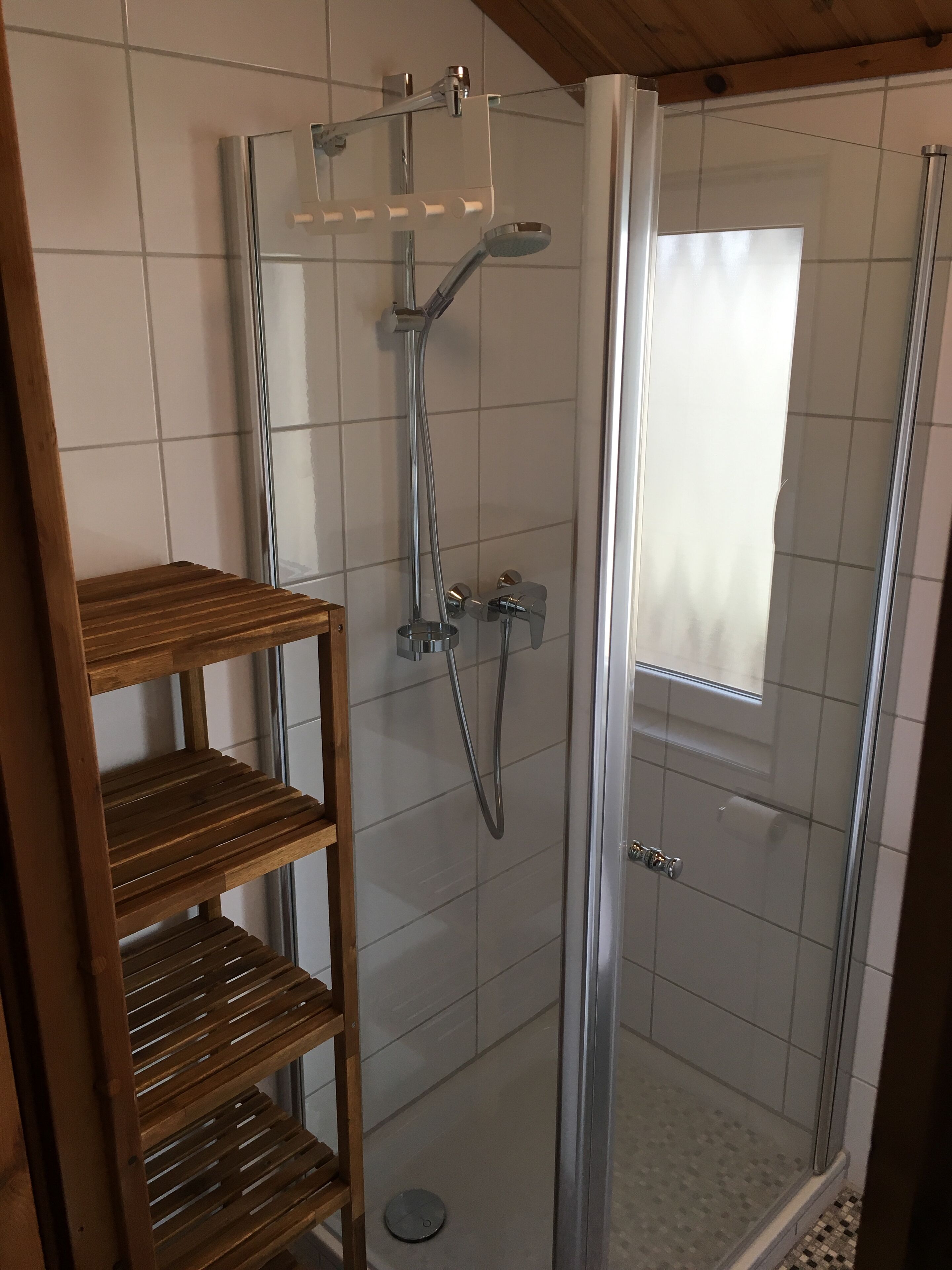 Cabina doccia con pareti in vetro, rubinetteria cromata e soffione manuale, un mobiletto a mensole in legno e una piccola finestra con tenda bianca.