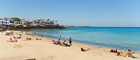 Une plage où l’on voit des gens se prélasser au soleil et nager, des bâtiments en arrière-plan et un ciel bleu dégagé.