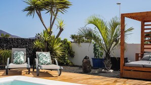 Un espace aménagé au bord de la piscine, avec un lit en bois, des chaises longues et des plantes tropicales.