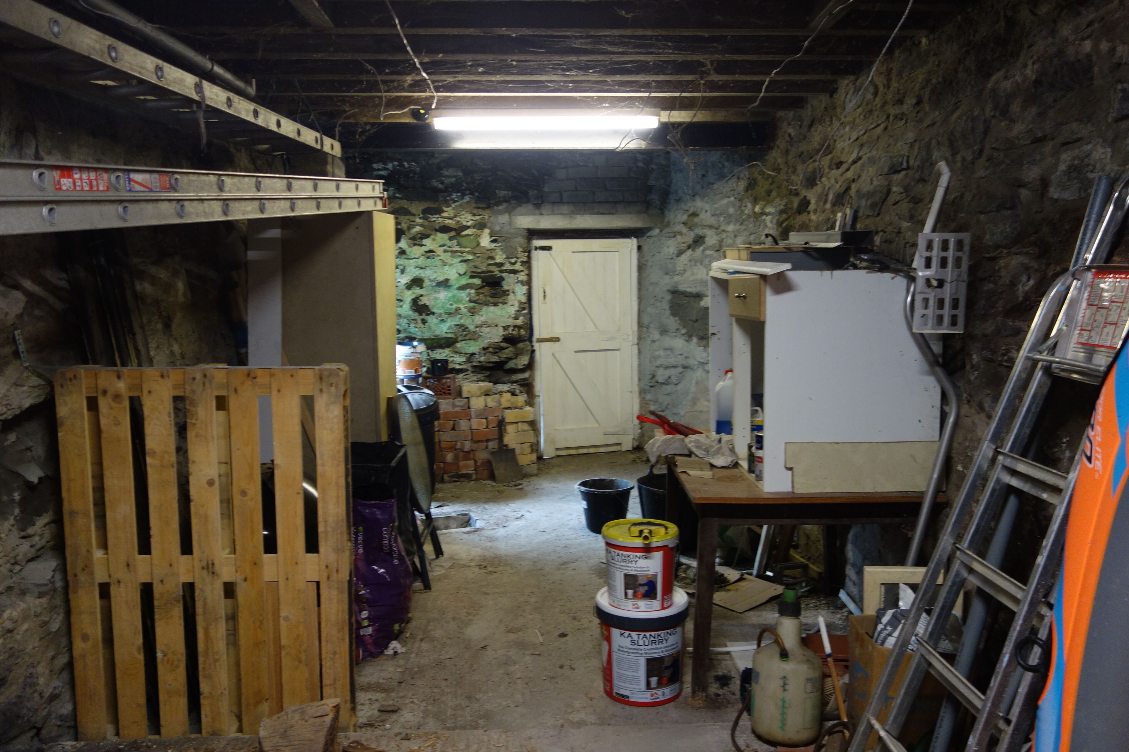 A narrow, dimly lit basement with a wooden pallet, a white door, and various construction materials.