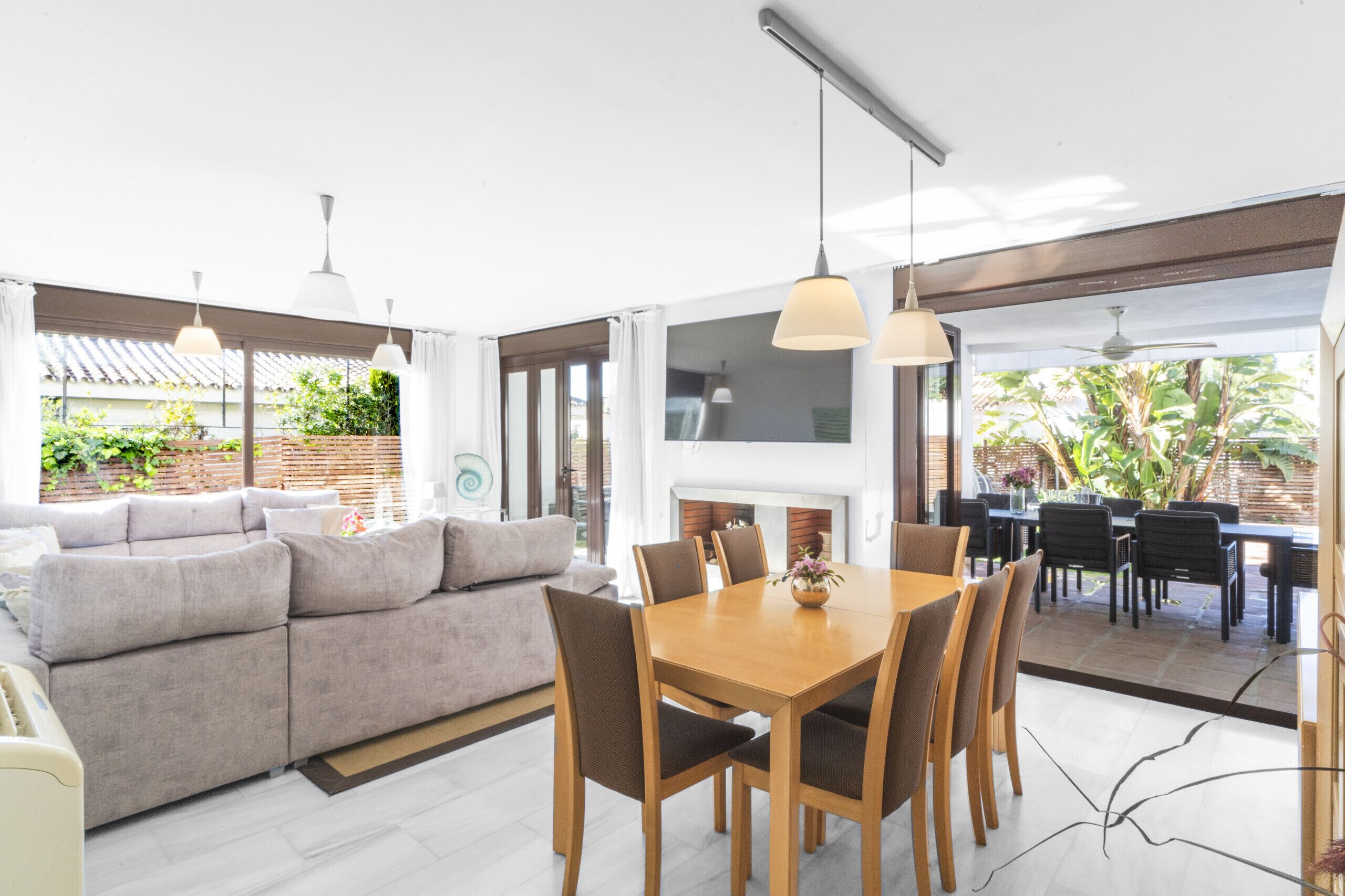 A modern living room with a sofa, dining table, and chairs. There is a fireplace and a view of the outdoors.