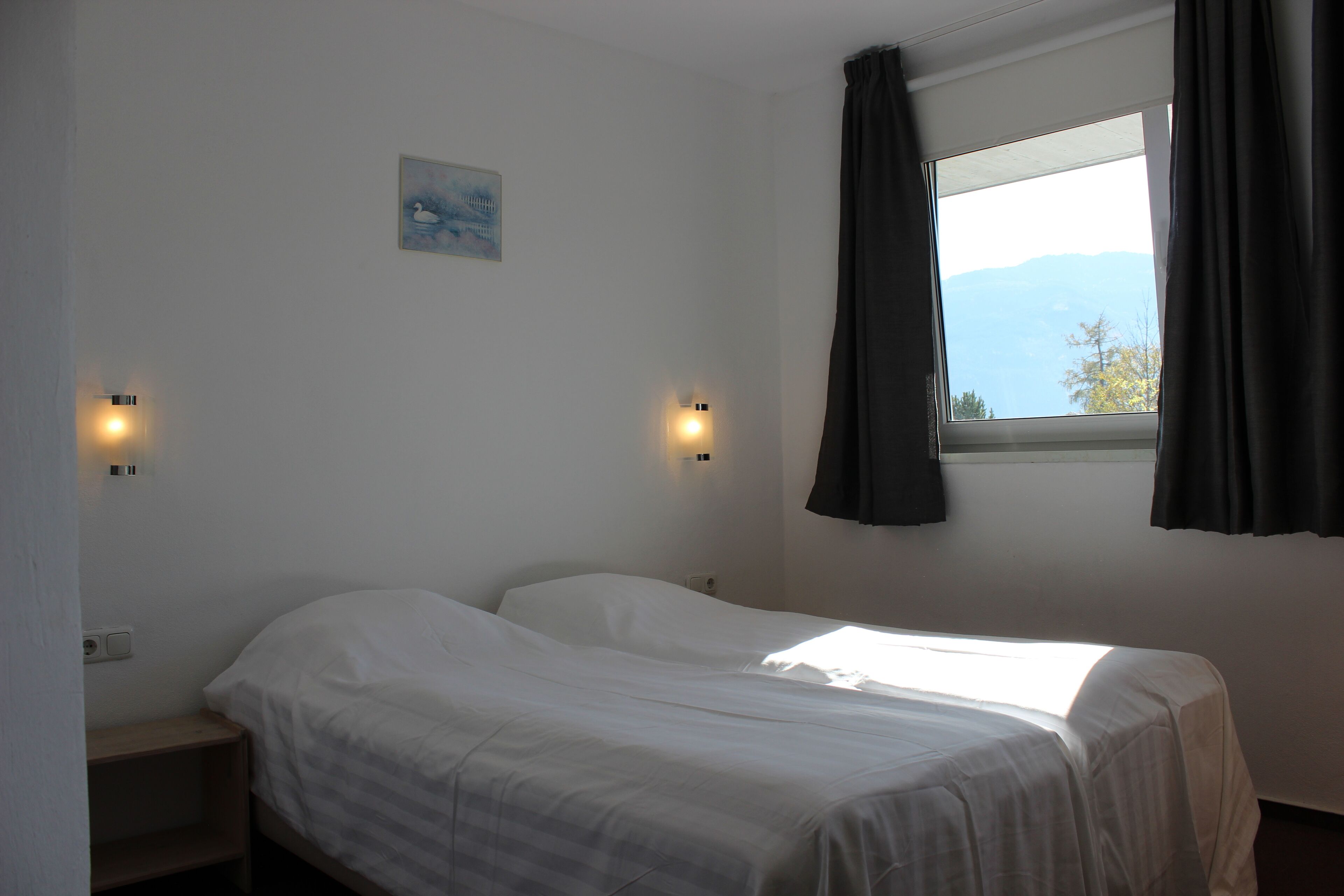 A single bed with white bedding, two wall-mounted light fixtures, a small painting on the wall, and a window with curtains.