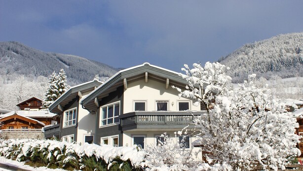 Um edifício grande de dois andares com uma varanda, rodeado por árvores cobertas de neve e montanhas ao fundo.