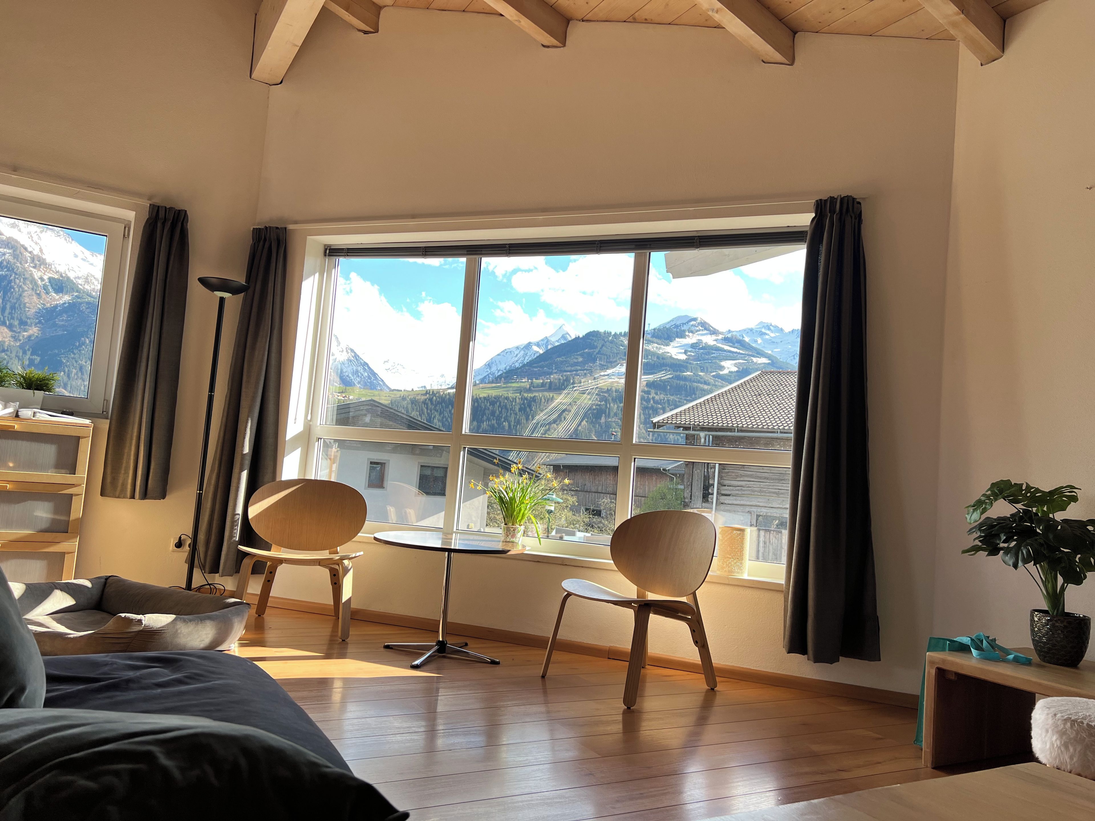 A room with a large window, wooden floor, a bed, two chairs, a small table, and a potted plant.