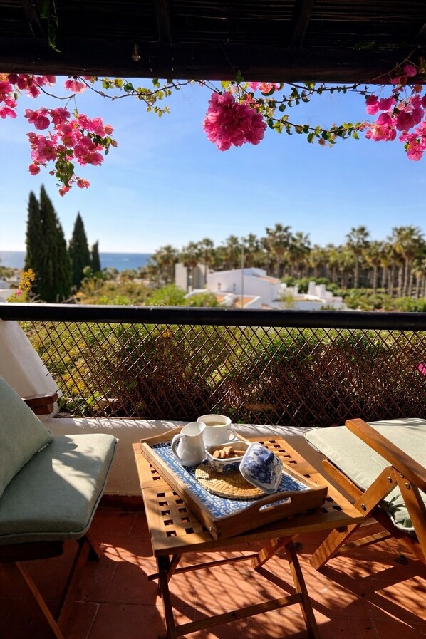 A terrace with a table set for two, a view of the sea, and a pergola with flowers.