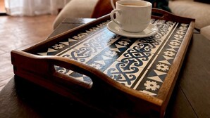A tray with a cup of coffee on a patterned table, a sofa, and a fireplace in a room with curtains and two framed pictures on the wall.