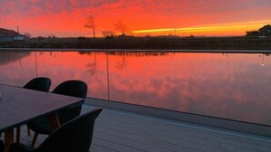 Auringonlasku tyynen vesistön yllä, etualalla puinen terassi ja tuoleja.