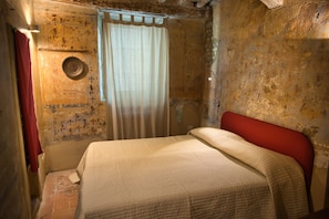 Une petite pièce rustique, avec un lit simple, une tête de lit rouge, un rideau et un luminaire fixé au mur.