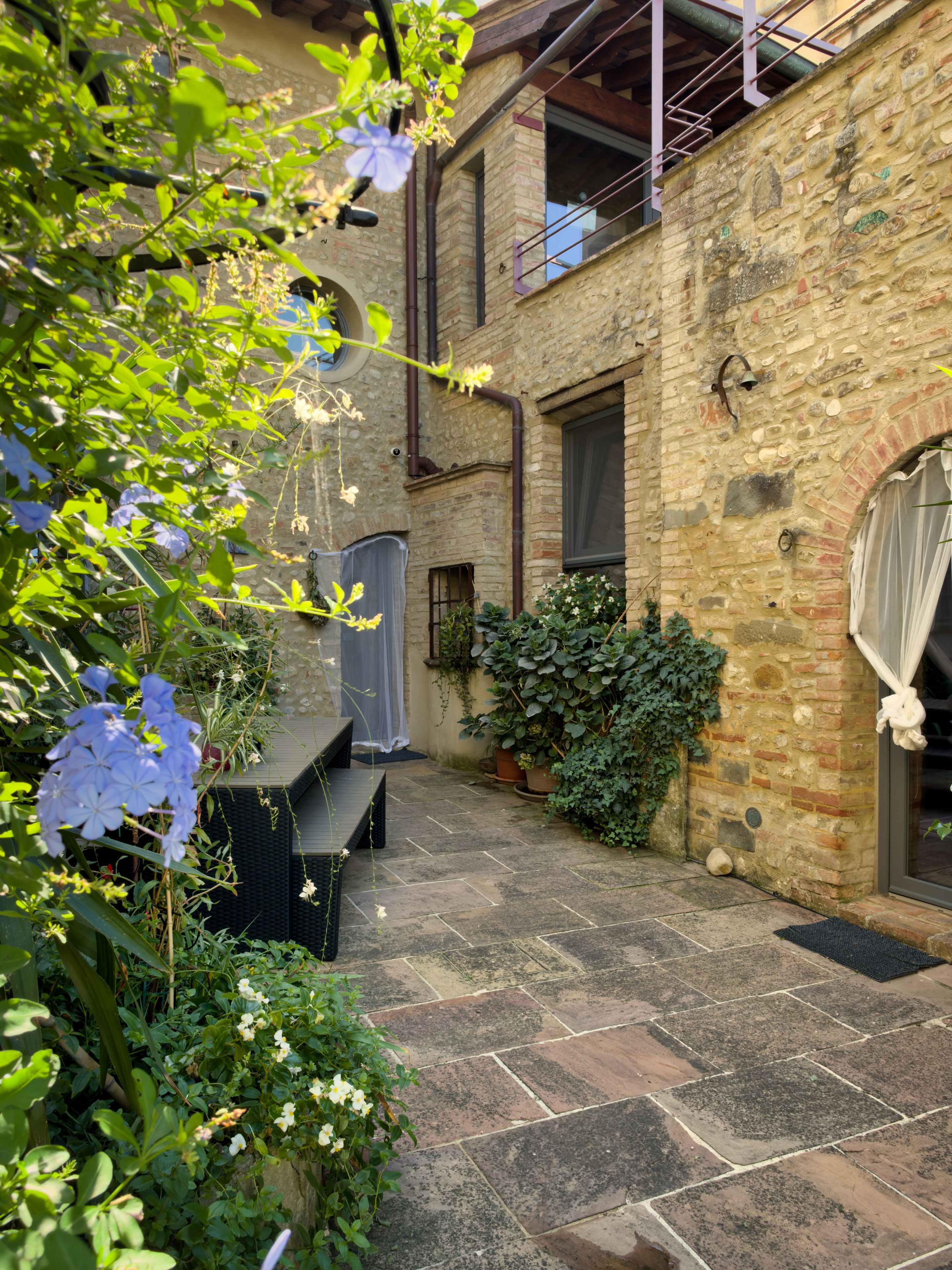 A courtyard with stone flooring, a wooden bench, potted plants, and a brick building with a window and a door.