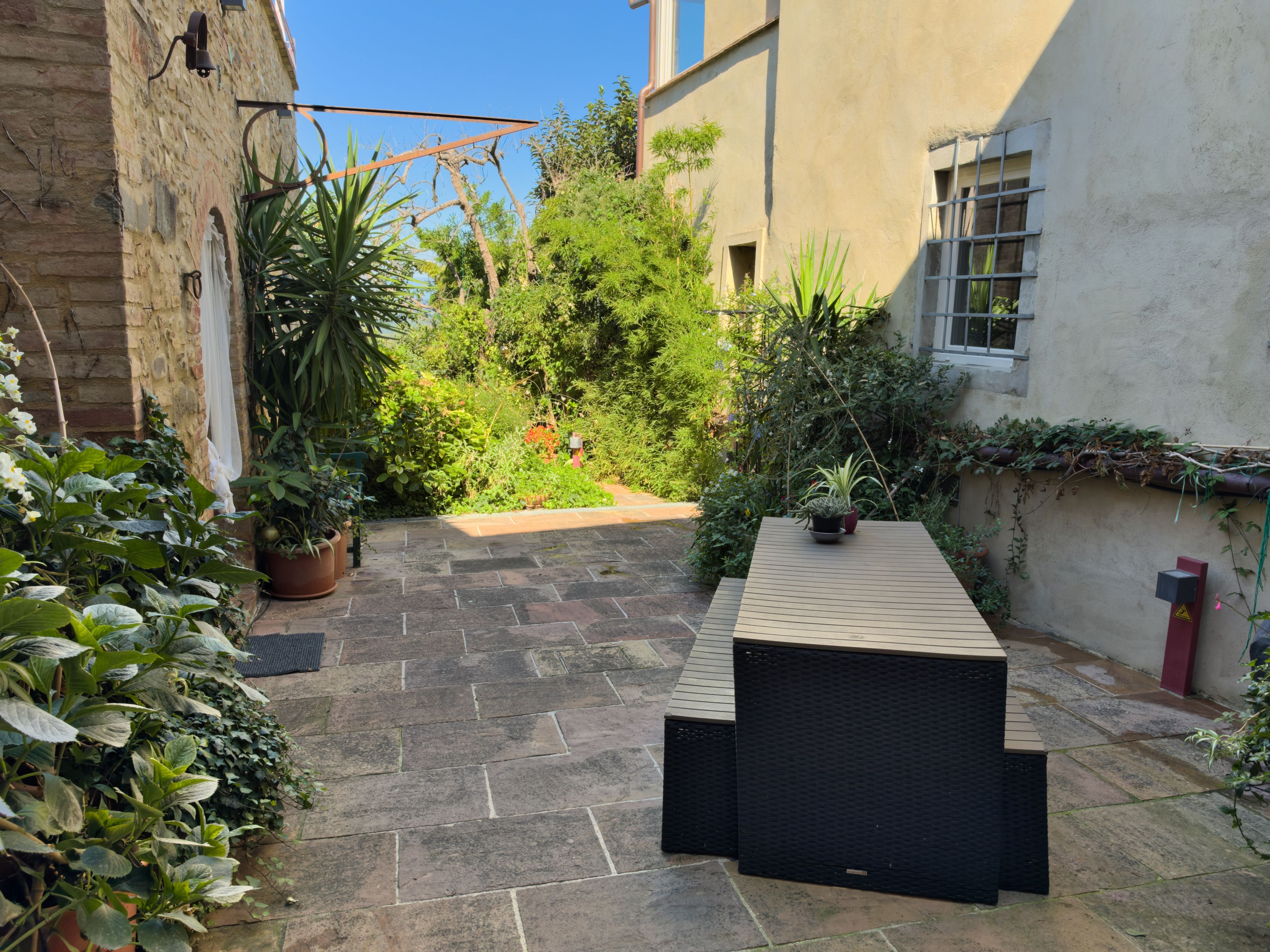 A courtyard with a stone floor, potted plants, a black outdoor table, and a building with a window.