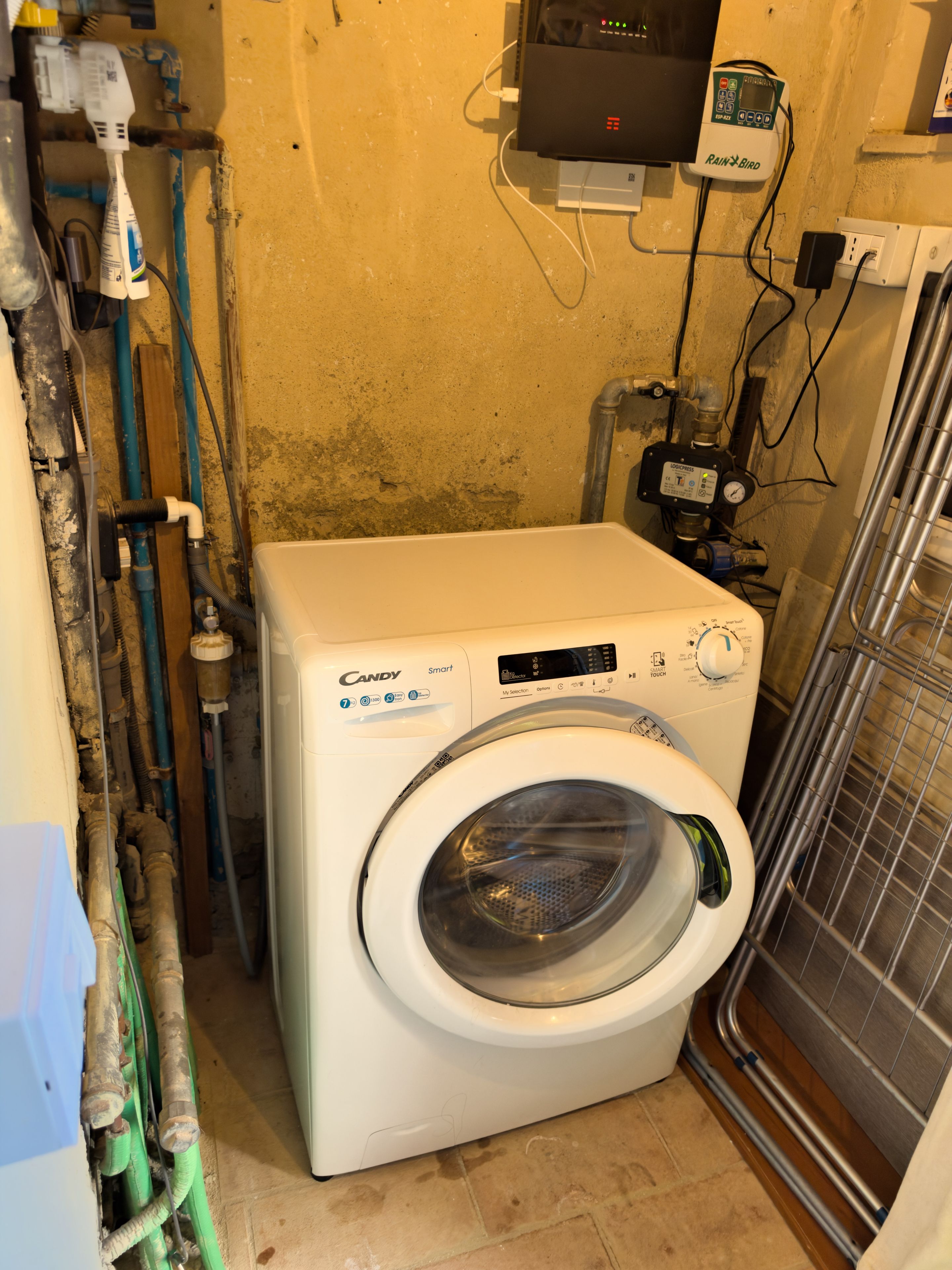 A compact, white Candy Smart washing machine in a small laundry room with visible plumbing and a water heater.