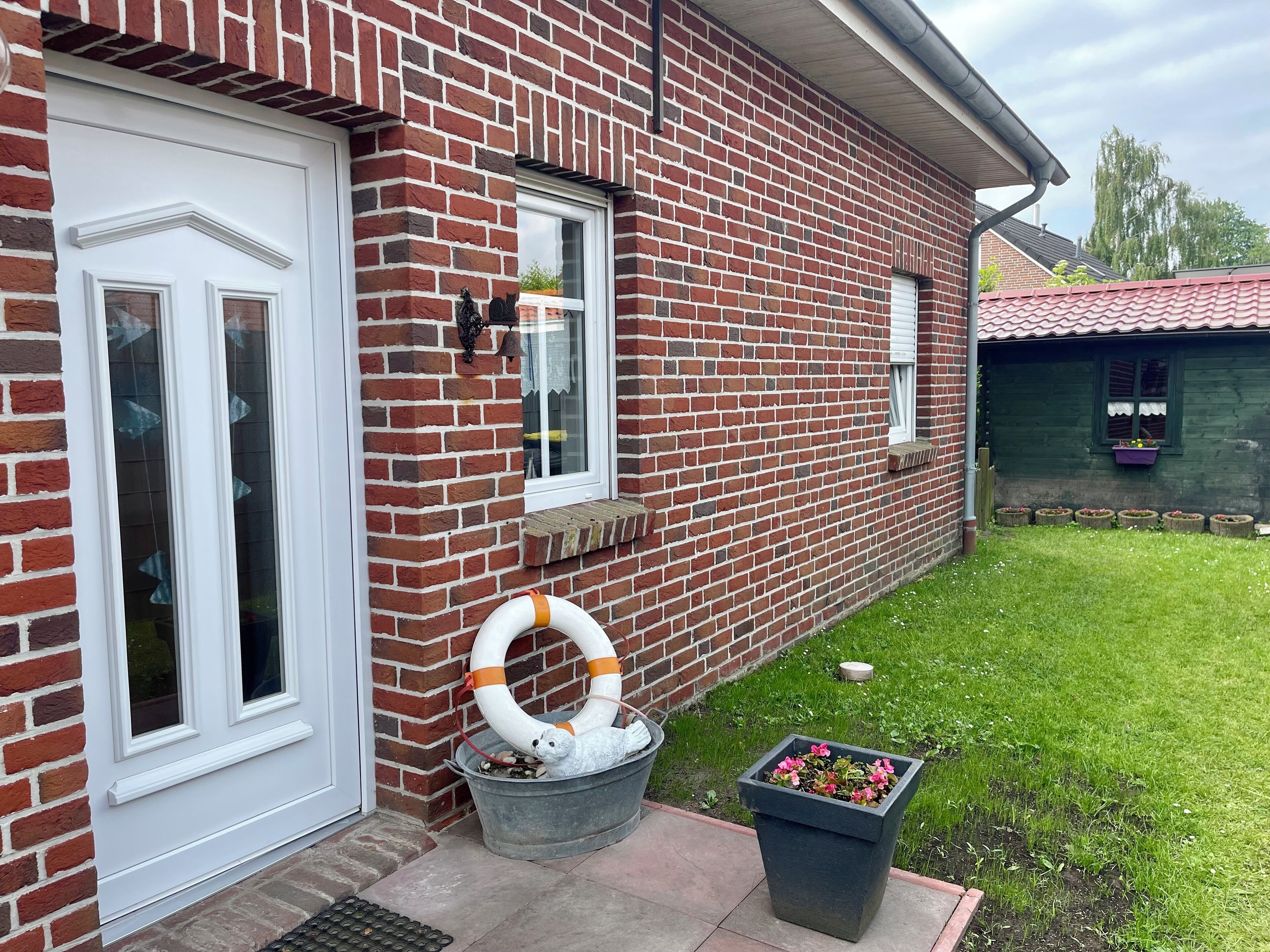 Ein Backsteinhaus mit einer weißen Tür, einem Fenster mit einer Klingel und einem Garten mit einem Blumentopf.