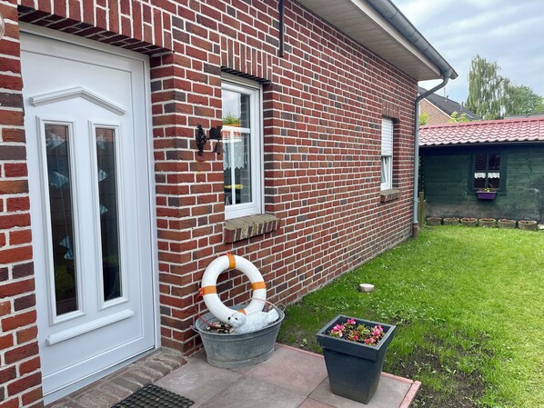 A brick house with a white door, a window with a bell, and a garden with a flower pot.