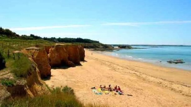 Una spiaggia con un gruppo di persone sedute sulla sabbia, scogliere sullo sfondo e cielo sereno.