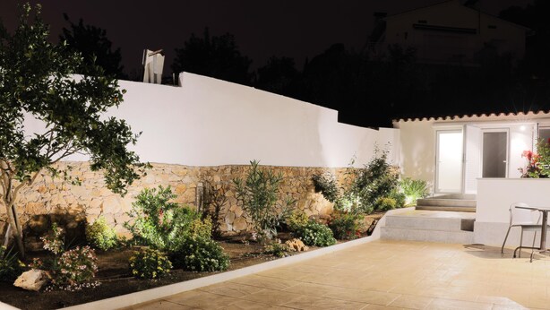 Un patio exterior cuidado con una pared de piedra, diversas plantas y una zona de estar con una silla y una mesa.