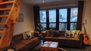 Ein gemütliches Wohnzimmer mit einem braunen Ledersofa, einem Holztisch und einer Gitarre. Durch das Fenster ist ein Weihnachtsbaum zu sehen.